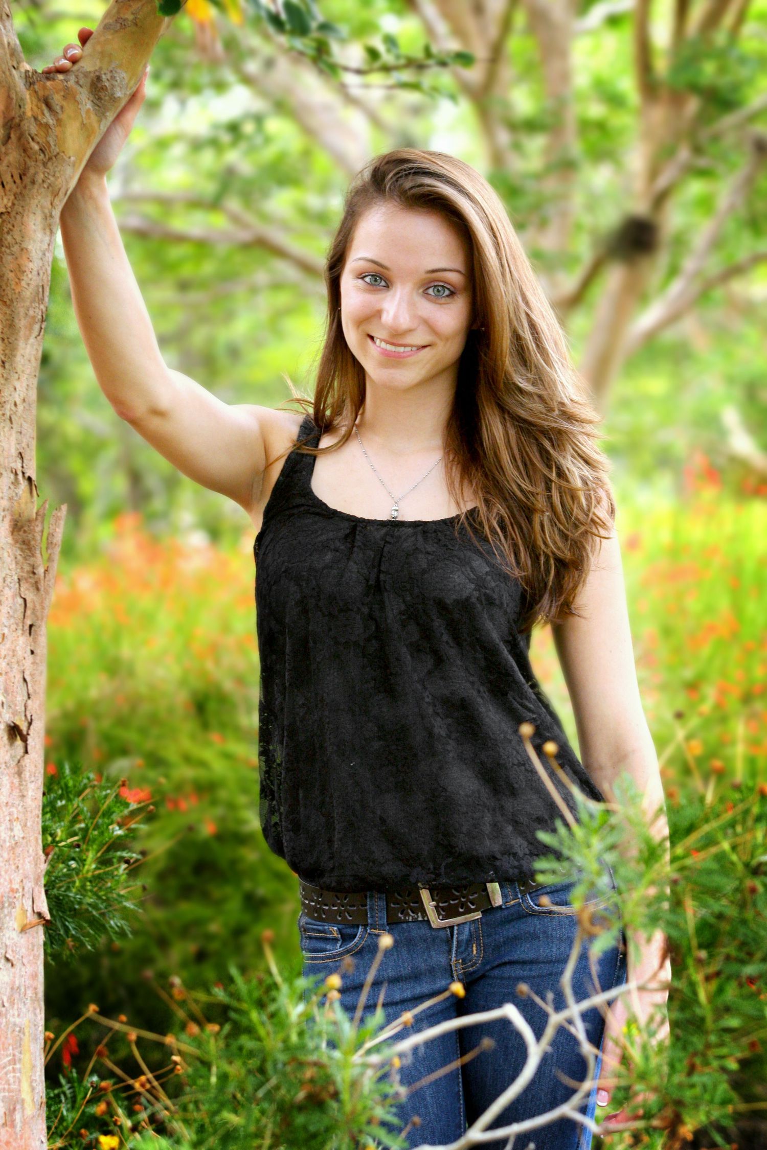Teenage girl in black shirt and blue jeans posing in a garden leaning on a tree.