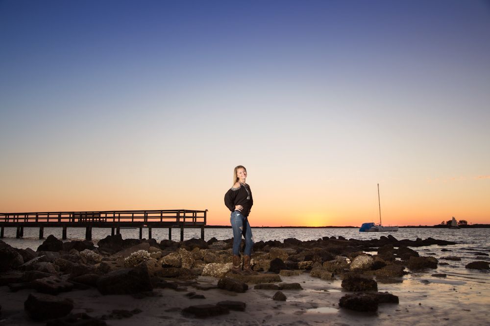 Girl in black top and blue jeans on rocky sea shore at golden sunset.
