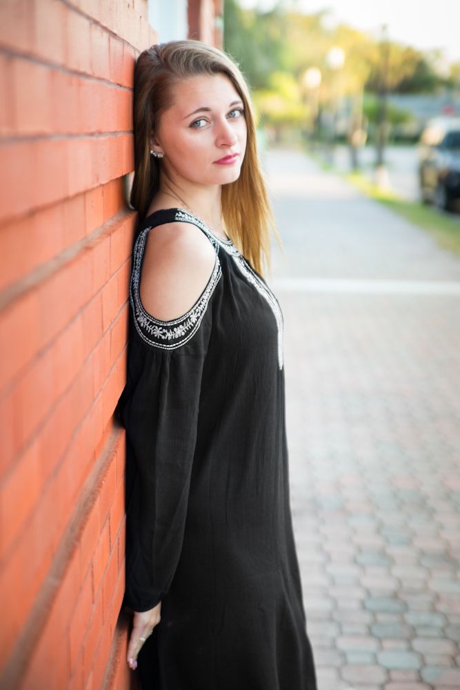 Teenage girl in black dress posing against brick wall.