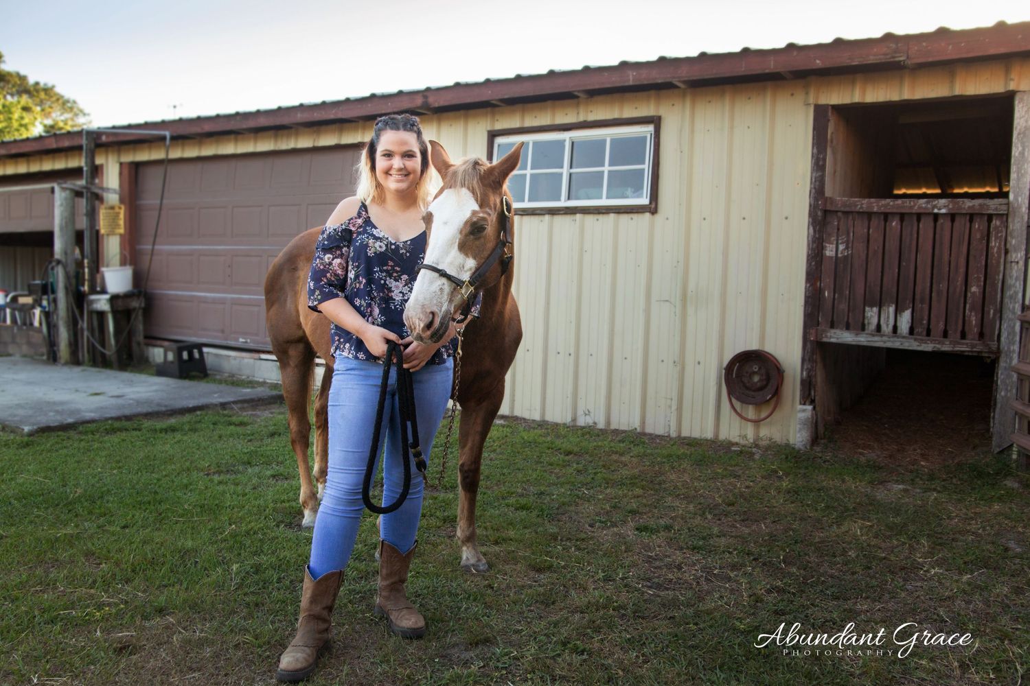 Girl in blue top and jeans standing with her brown horse at her barn.