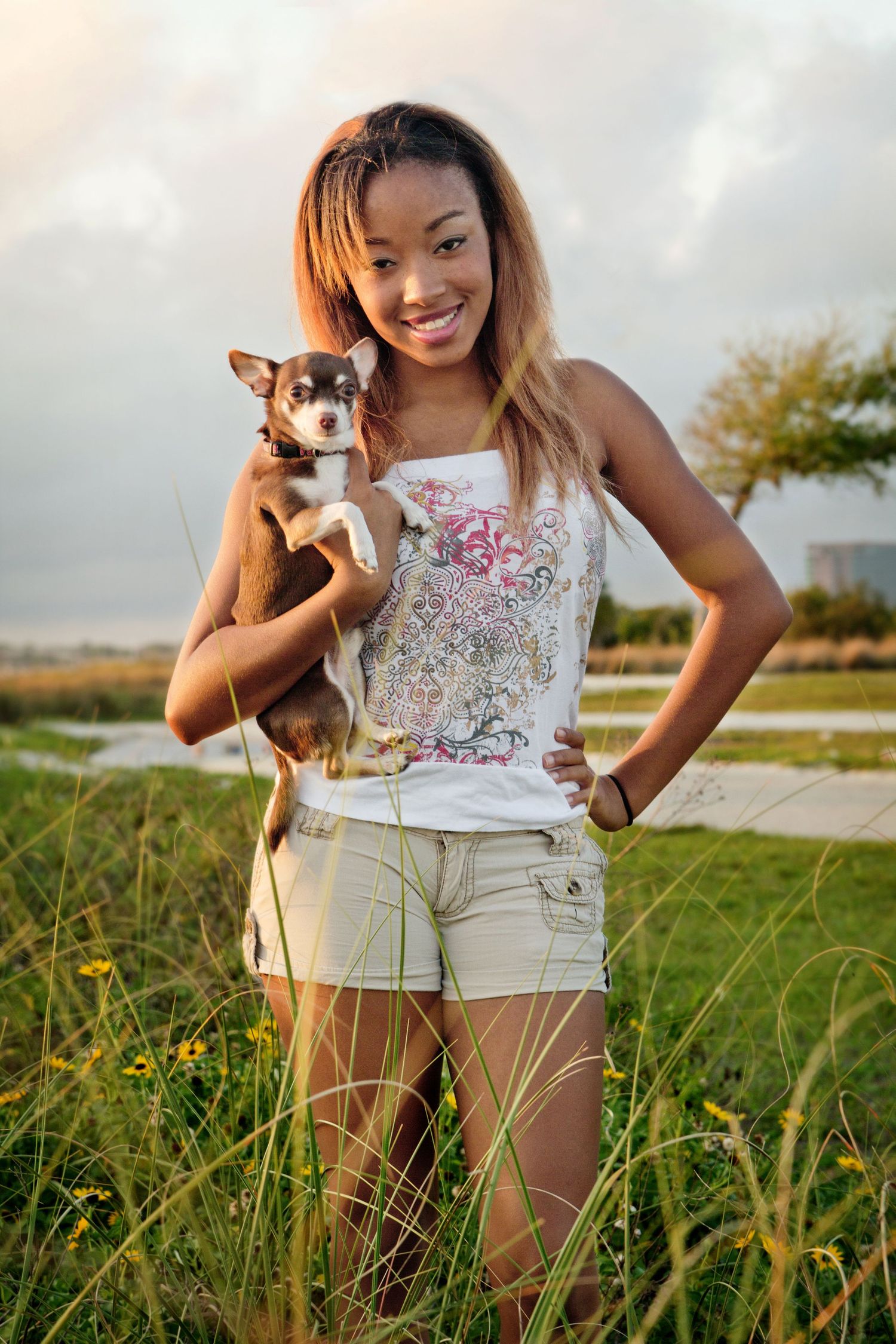 Teenage girl in white top and tan shorts posing with her small brown dog standing in the tall grass at Cypress Point Beach Park.