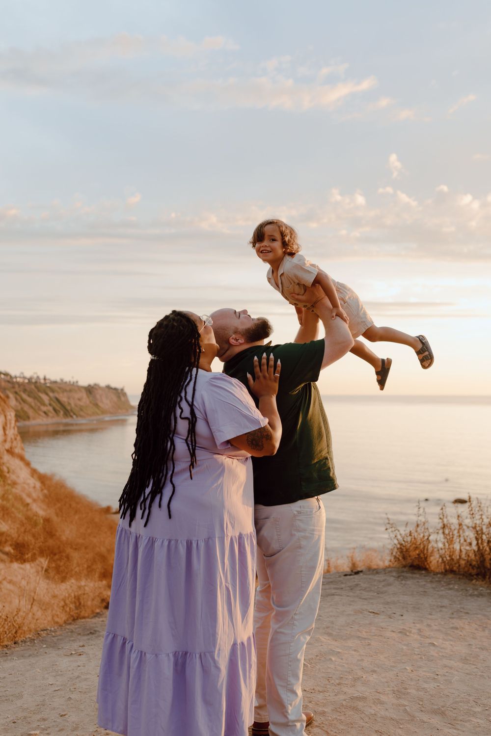 Family enjoying sunset moment on coastal cliff with child being lifted playfully.