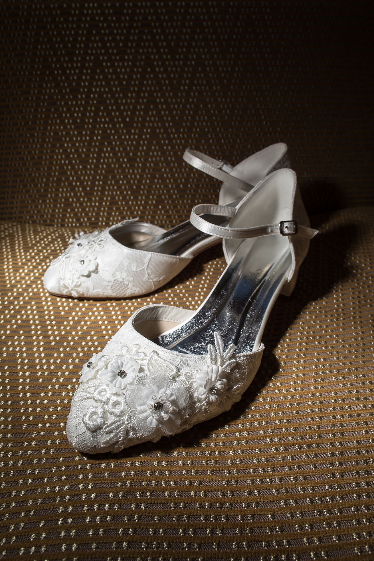 Vintage-style white lace wedding shoes with ankle straps on textured golden fabric.