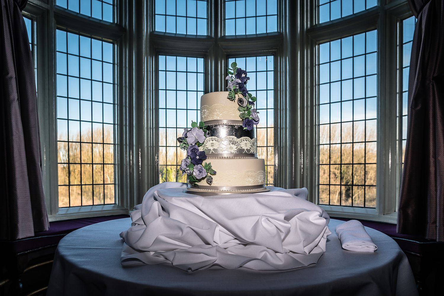 Three-tier white wedding cake displayed in bay window with scenic outdoor view.