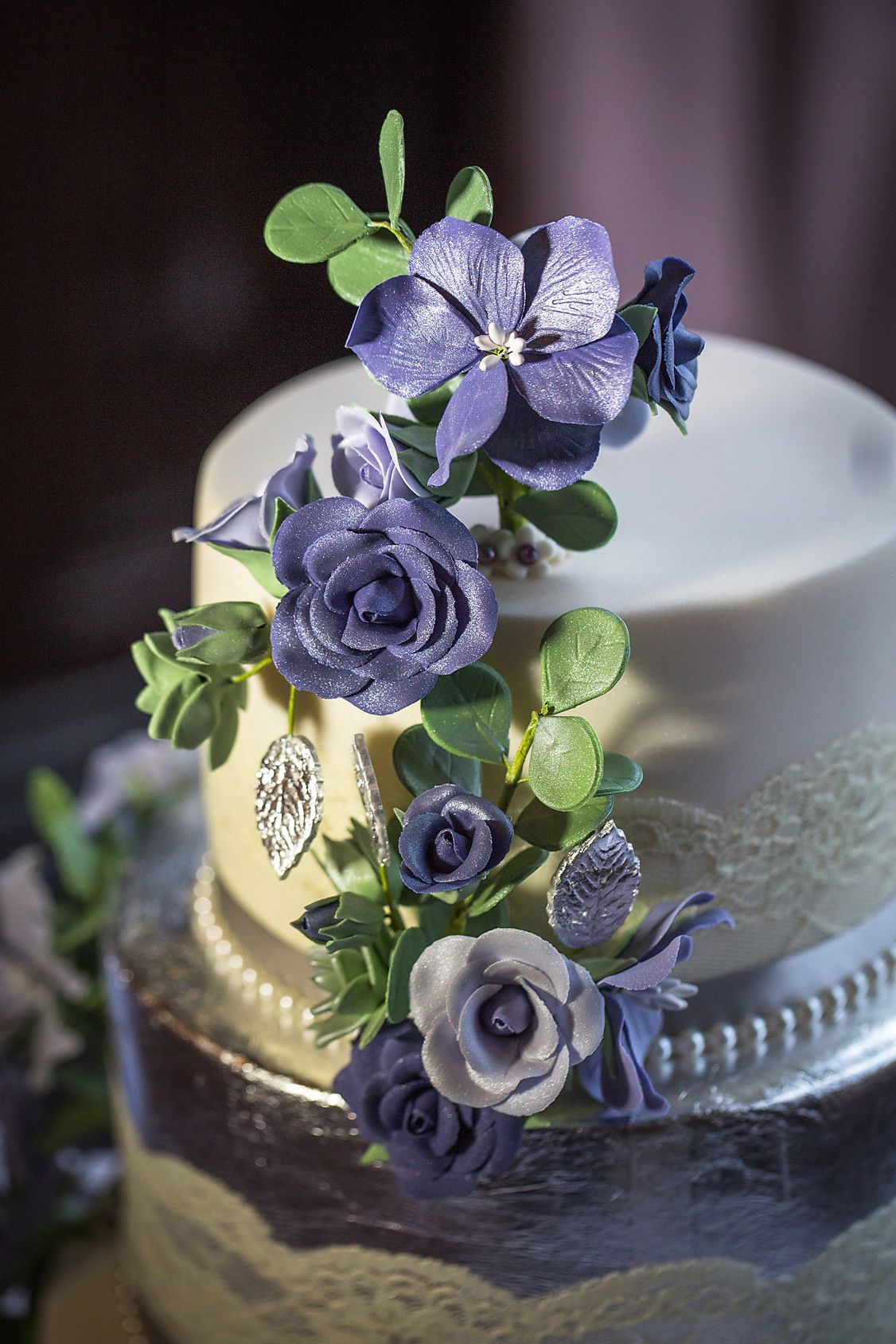 Elegant wedding cake detail with purple sugar flowers and green foliage decorations.