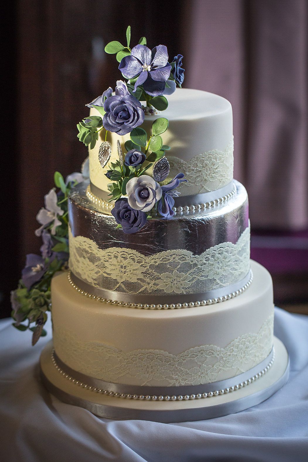Three-tier white wedding cake decorated with purple sugar flowers, lace details, and silver trim.