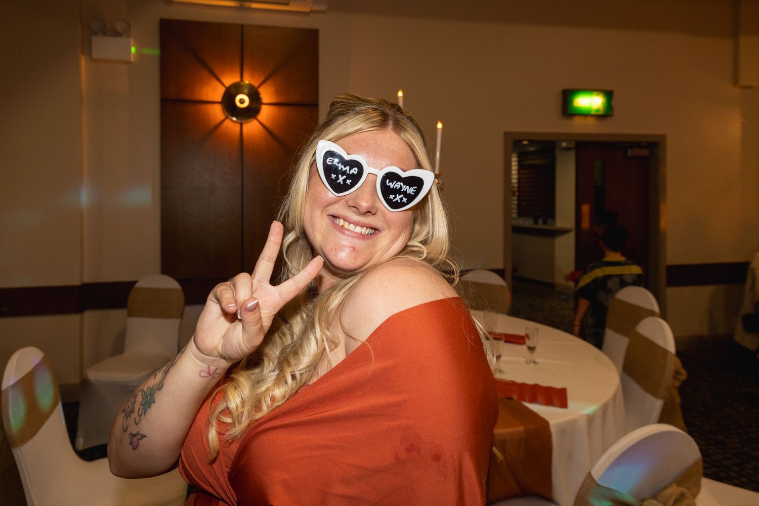 Person in orange top wearing heart sunglasses makes peace sign gesture at a banquet hall.