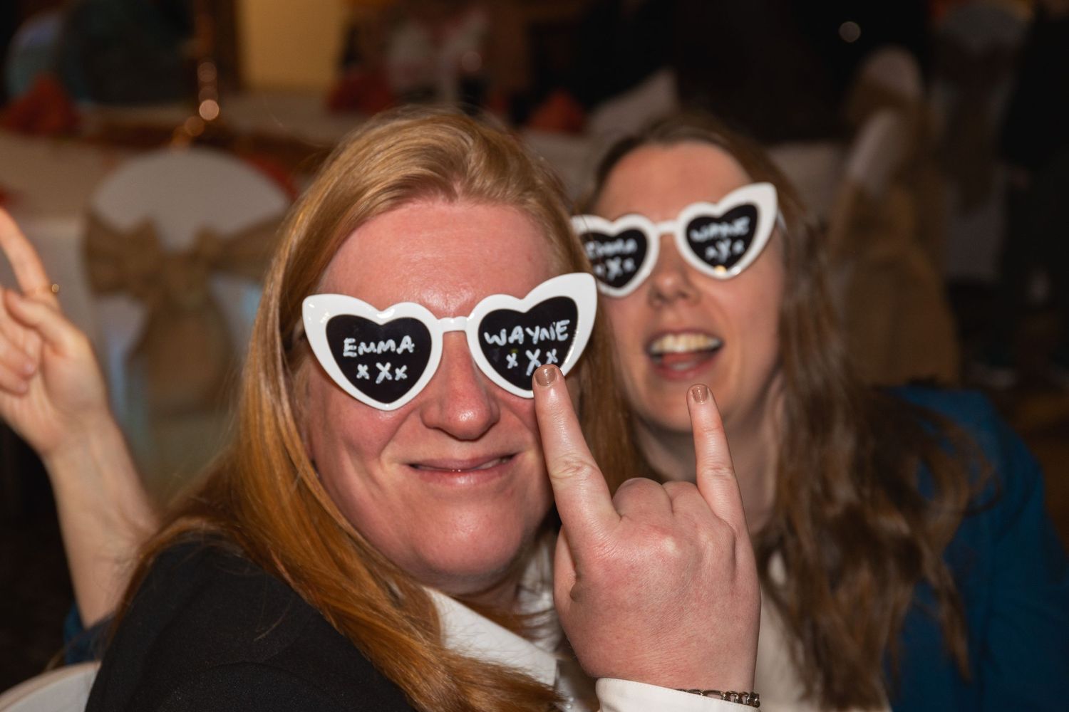 Person with long hair wearing heart-shaped glasses makes peace sign at celebration.