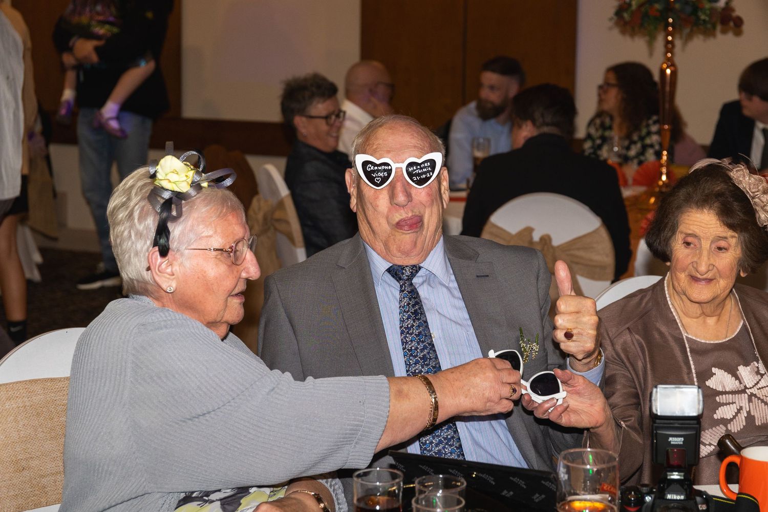 Three guests at dinner table, with one wearing heart-shaped sunglasses making animated gesture.