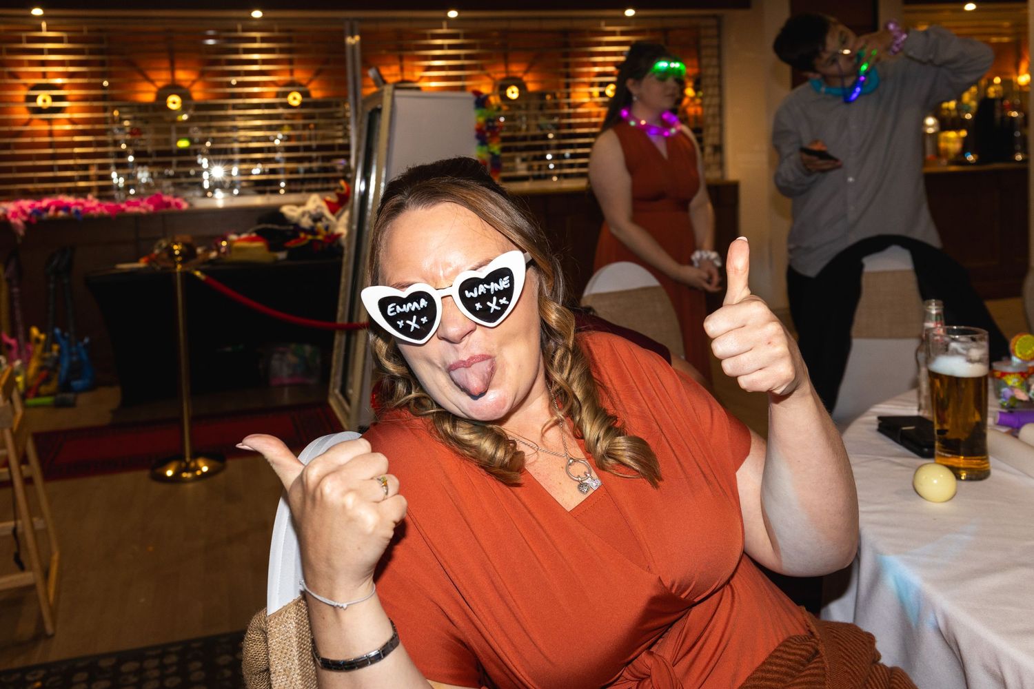 Person in orange top wearing heart sunglasses gives thumbs up while sitting at event.