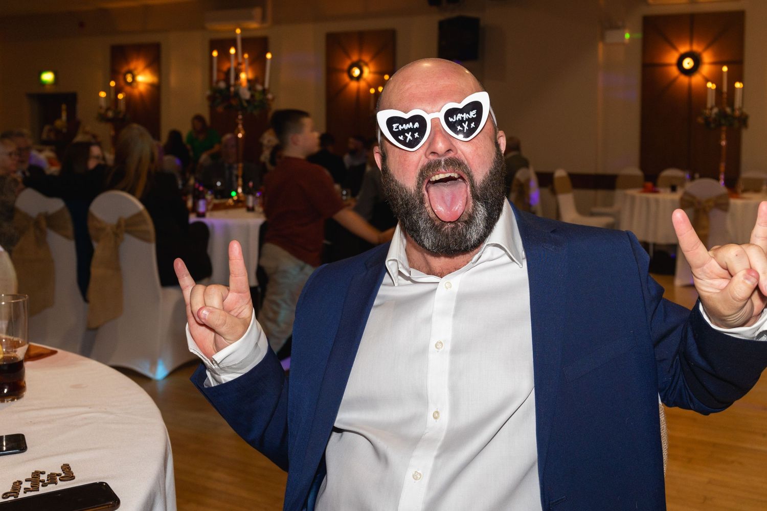 Person in blue suit and white shirt makes peace signs while wearing heart-shaped sunglasses at formal event.