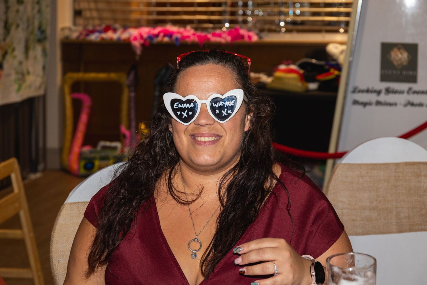 Person in burgundy attire wearing decorative heart sunglasses poses at indoor celebration.