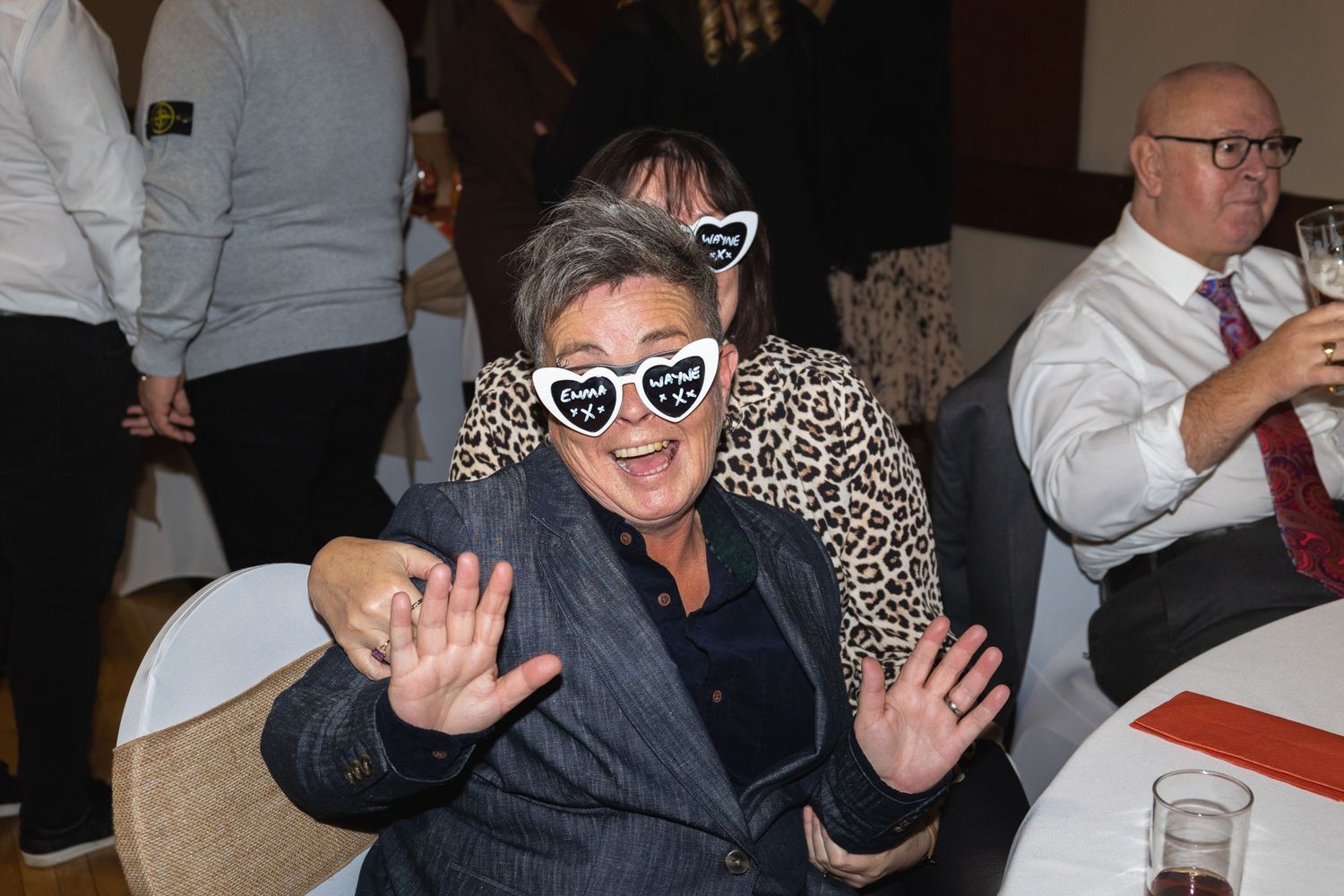 Guest making playful gesture while wearing heart-shaped glasses at formal celebration.