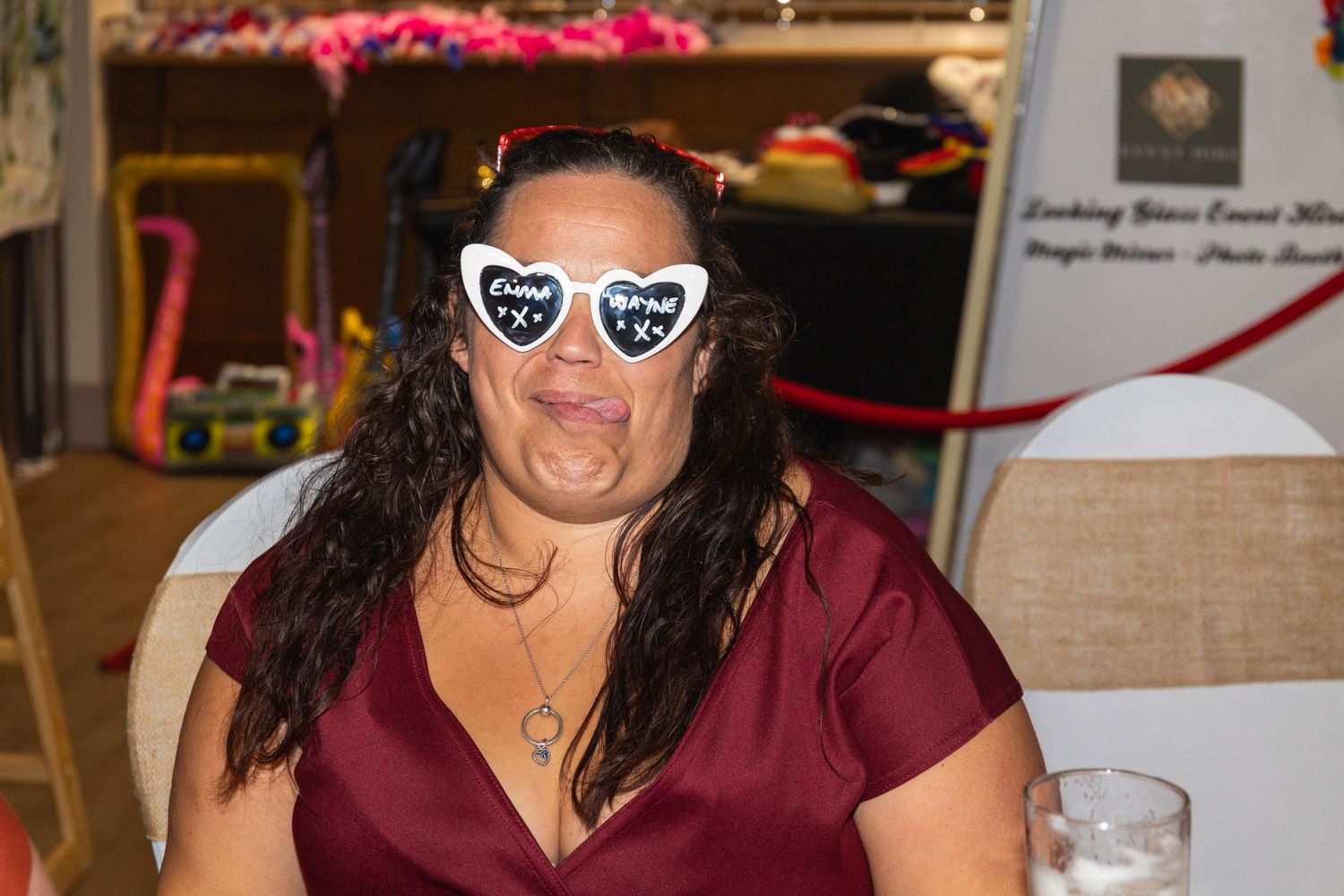 Person in burgundy top wearing heart-shaped sunglasses smiles enthusiastically at indoor gathering.