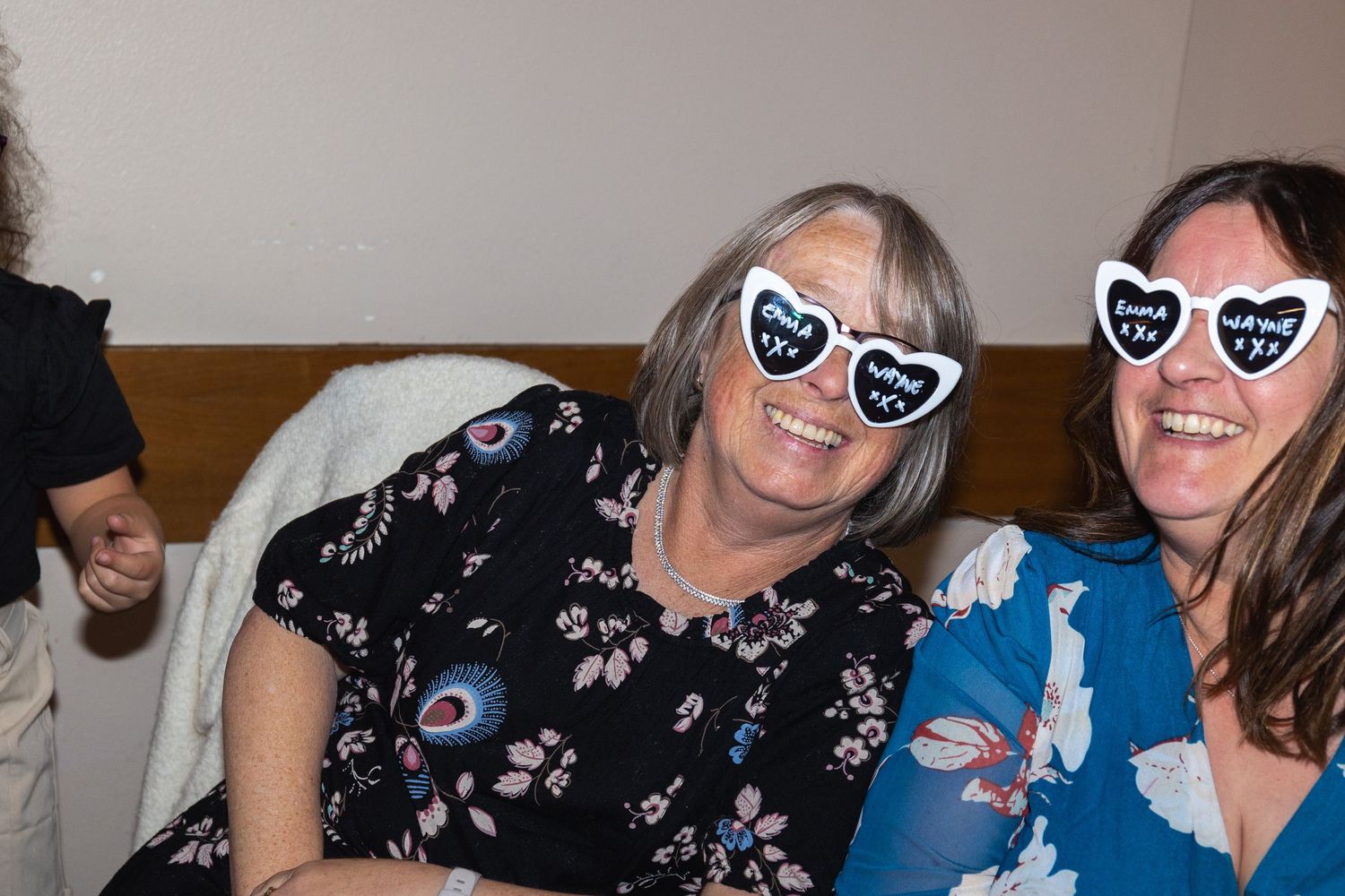 Two people wearing matching heart-shaped novelty sunglasses smile while sitting together at an event.