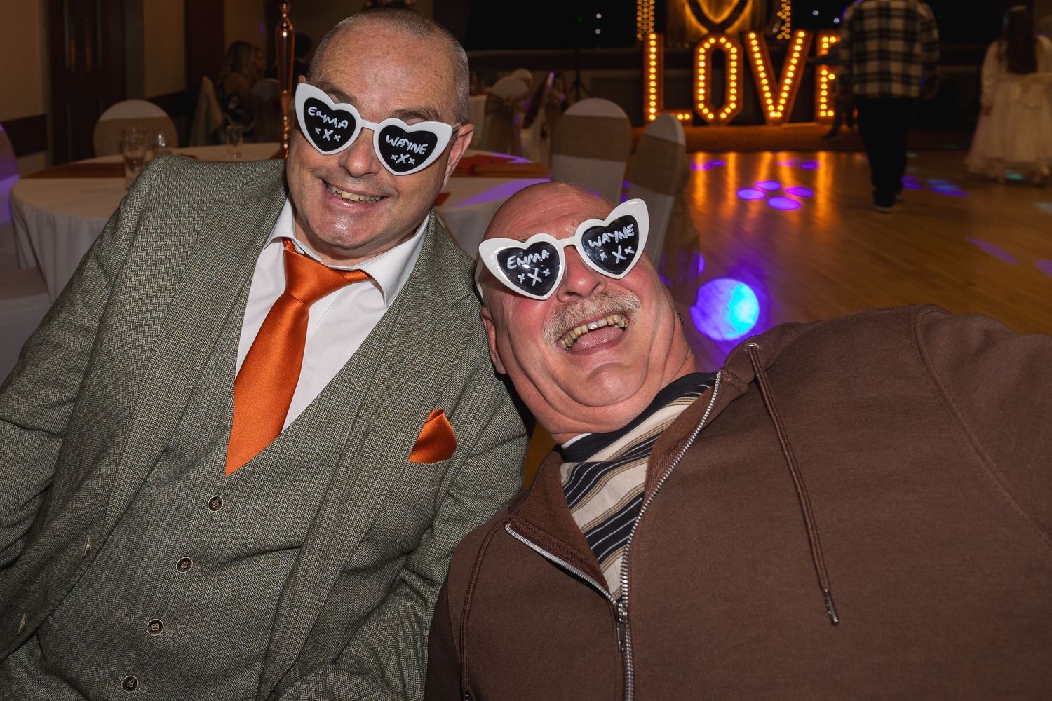 Two guests wearing matching heart glasses pose together at formal event with LOVE sign in background.
