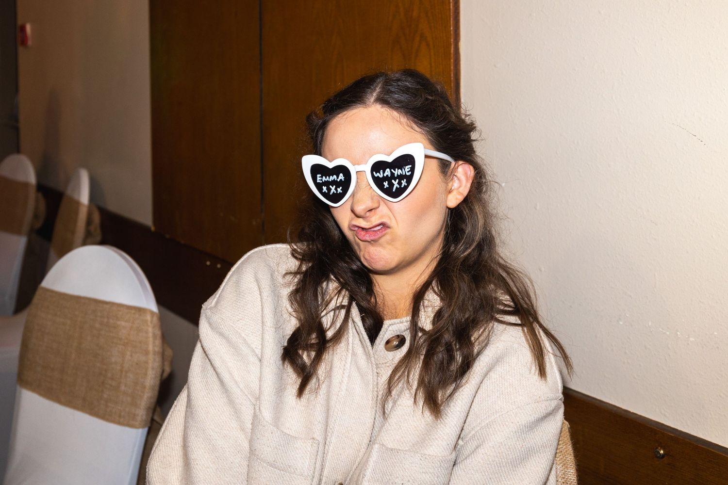 Person in beige outfit wearing heart-shaped sunglasses sits at indoor gathering.