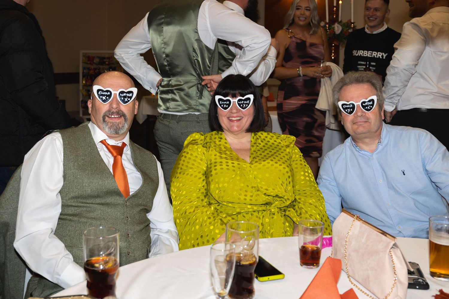 Three guests at a celebration wearing matching heart-shaped glasses while seated at a table.