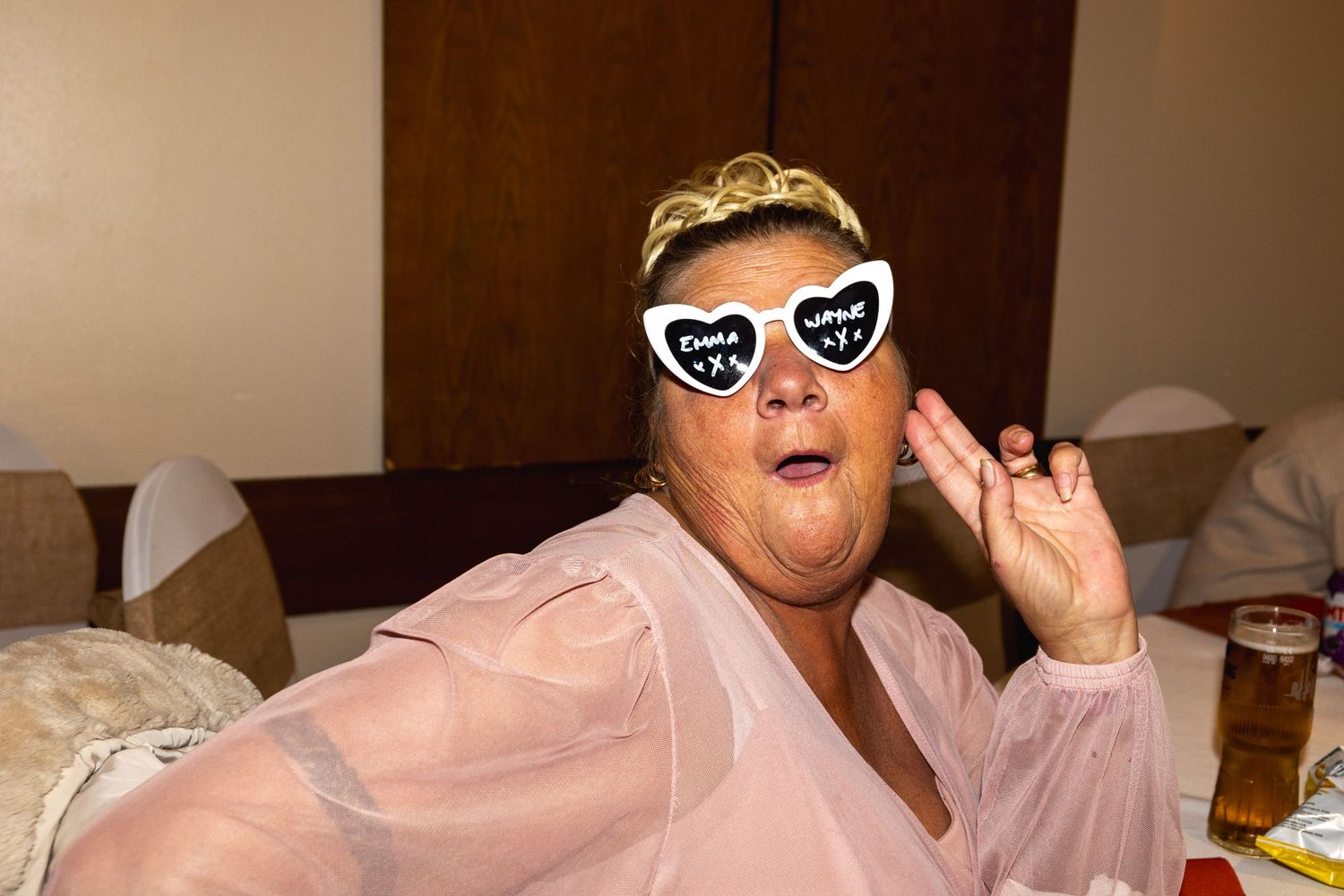 Person in pink robe wearing heart sunglasses and tiara poses on couch with beverage nearby.