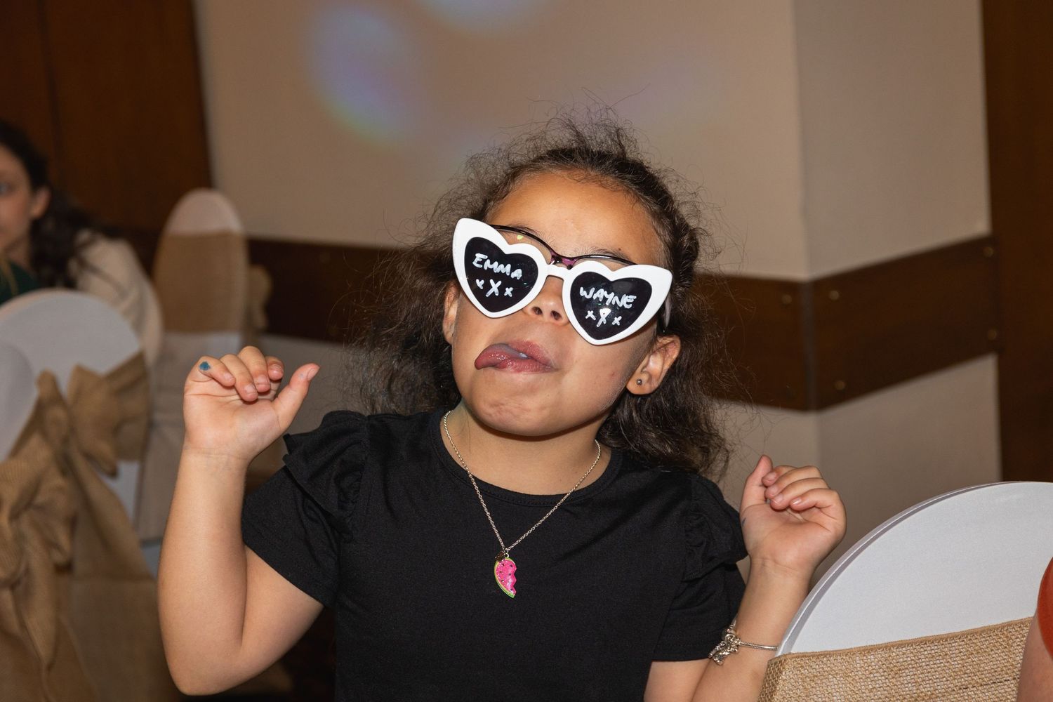 Person in black shirt wearing heart-shaped sunglasses poses with raised hands at indoor event.