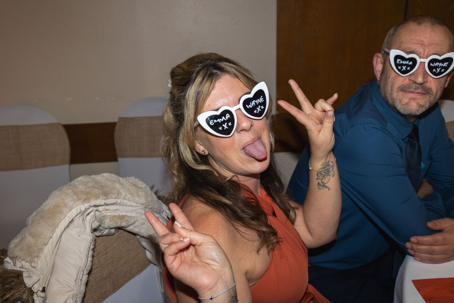 Two people wearing matching heart-shaped sunglasses making peace signs at a party.