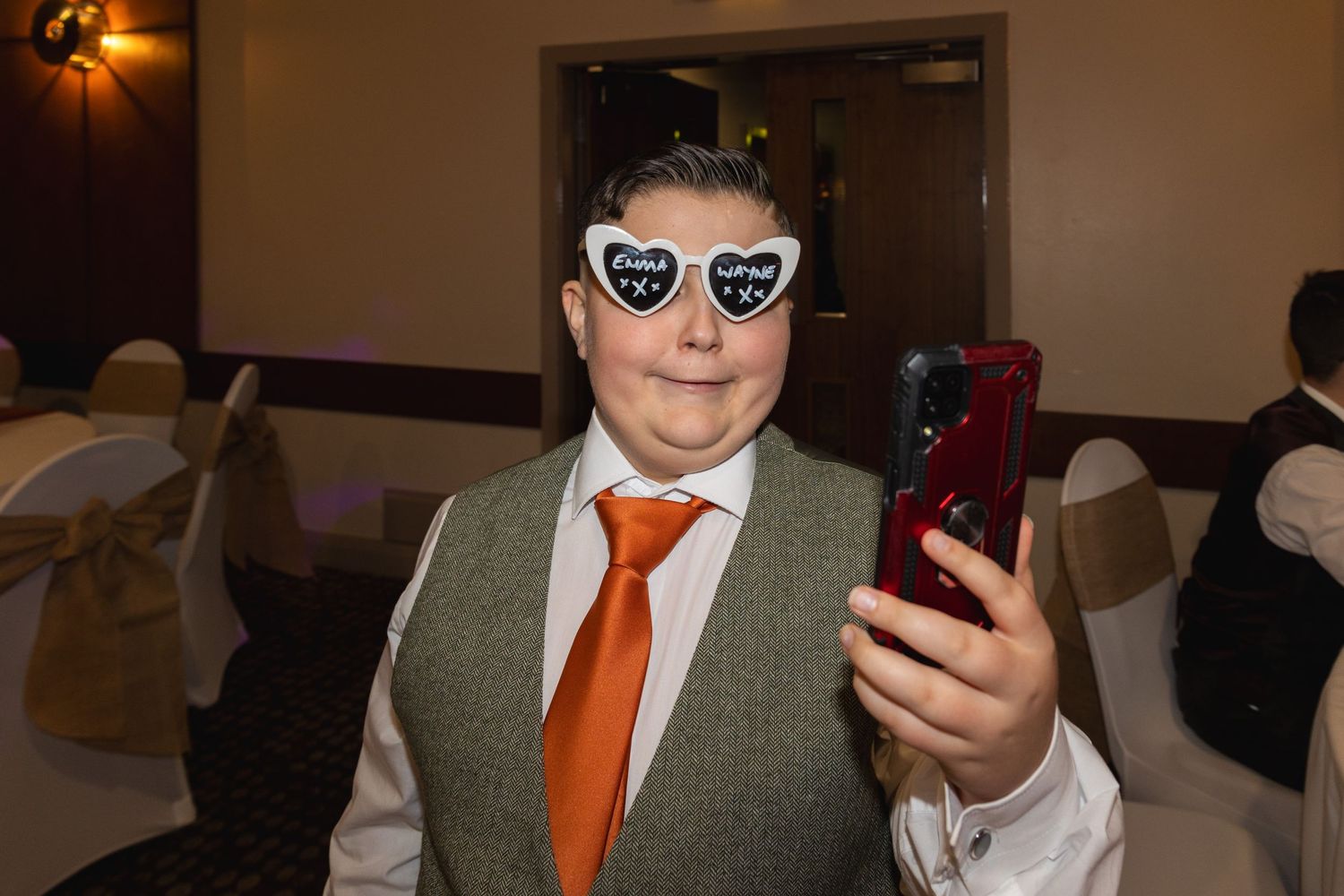 Event guest in grey vest and orange tie taking a selfie while wearing decorative glasses.