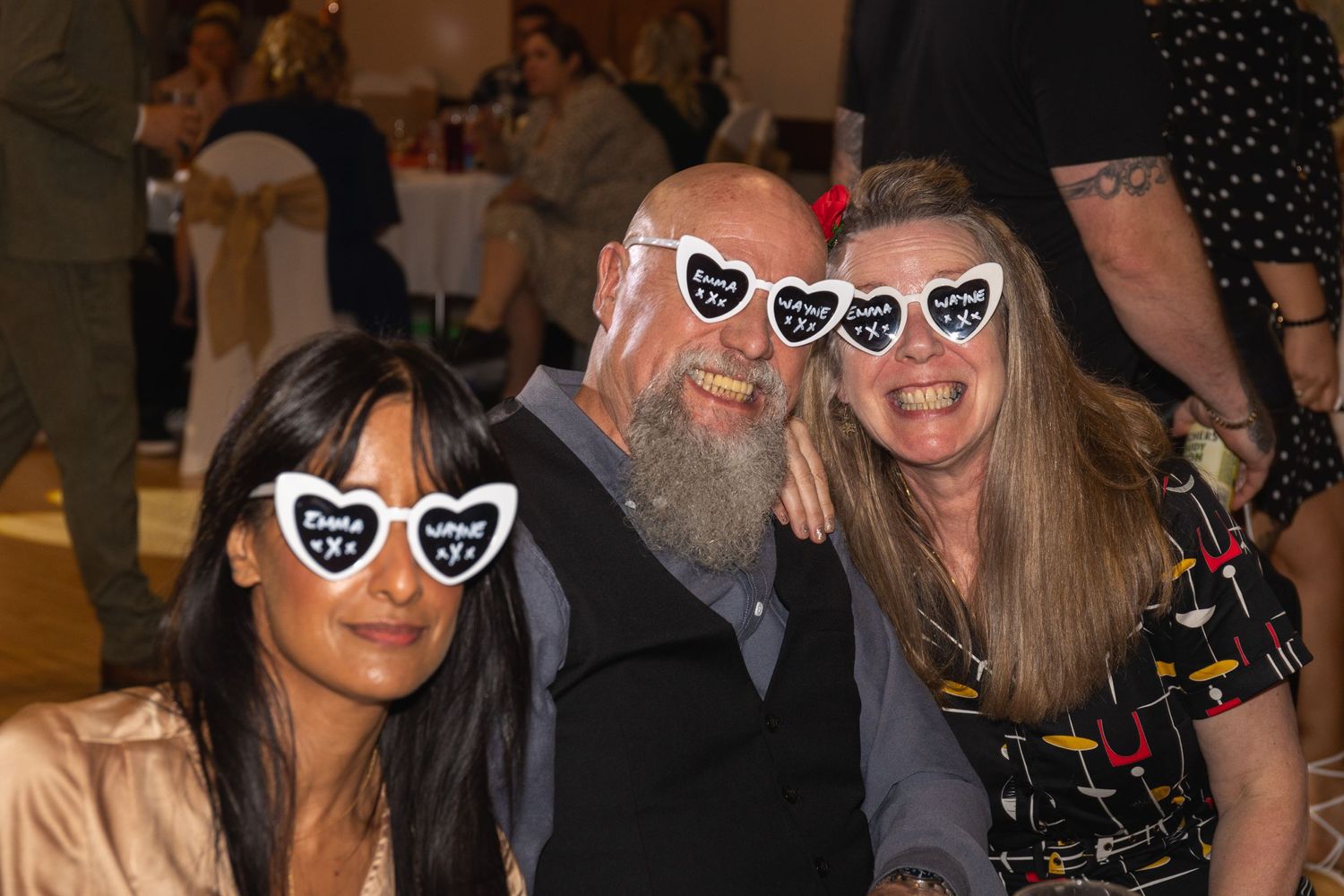 Three guests wearing matching heart-shaped sunglasses pose together at formal gathering.