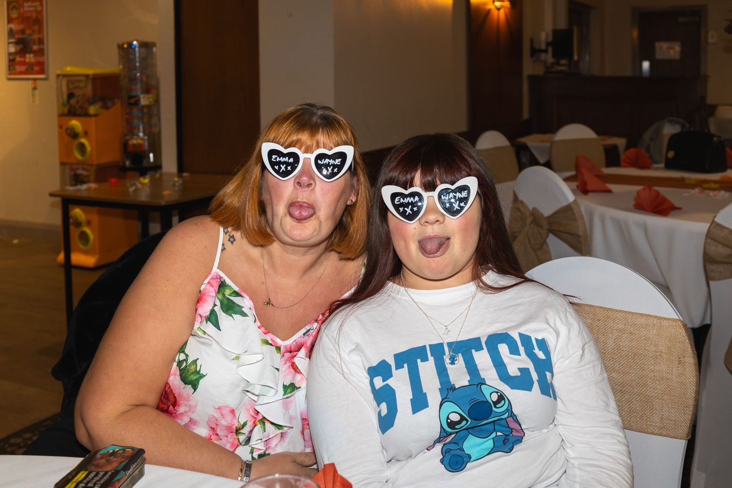 Two people wearing heart-shaped sunglasses pose together at an indoor event, one in floral print and one in Stitch shirt.