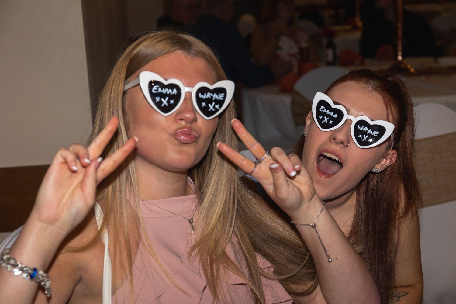 Two people in heart sunglasses make peace signs while posing together at an indoor venue.