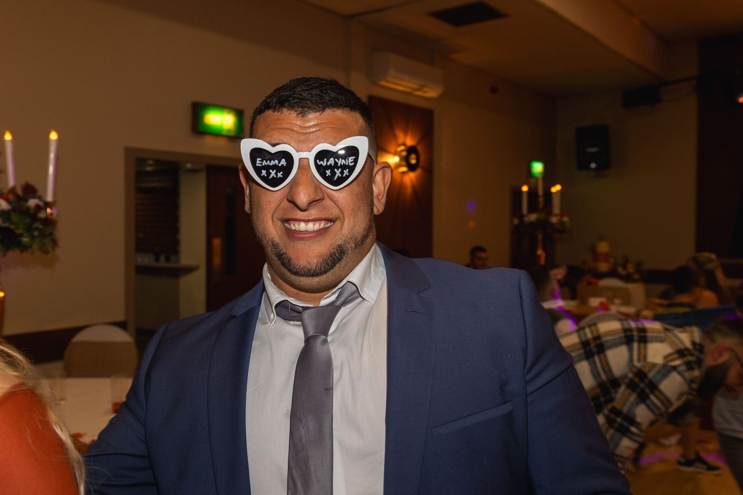 Smiling guest in blue suit and tie wearing heart-shaped sunglasses at formal event.
