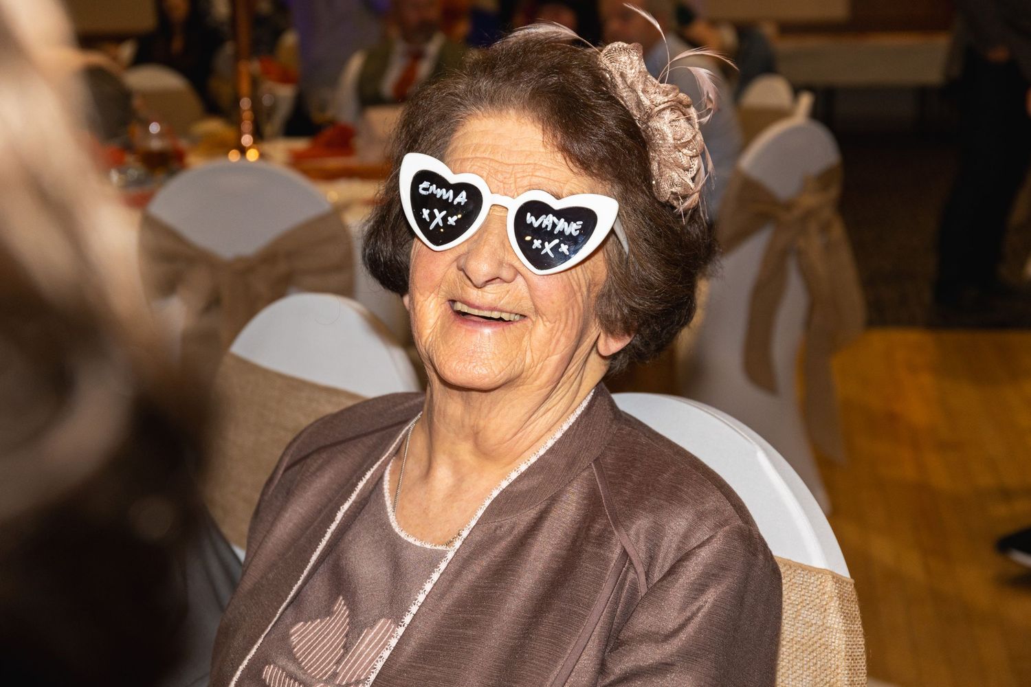 Senior woman in brown jacket wearing heart-shaped novelty glasses smiles at party.