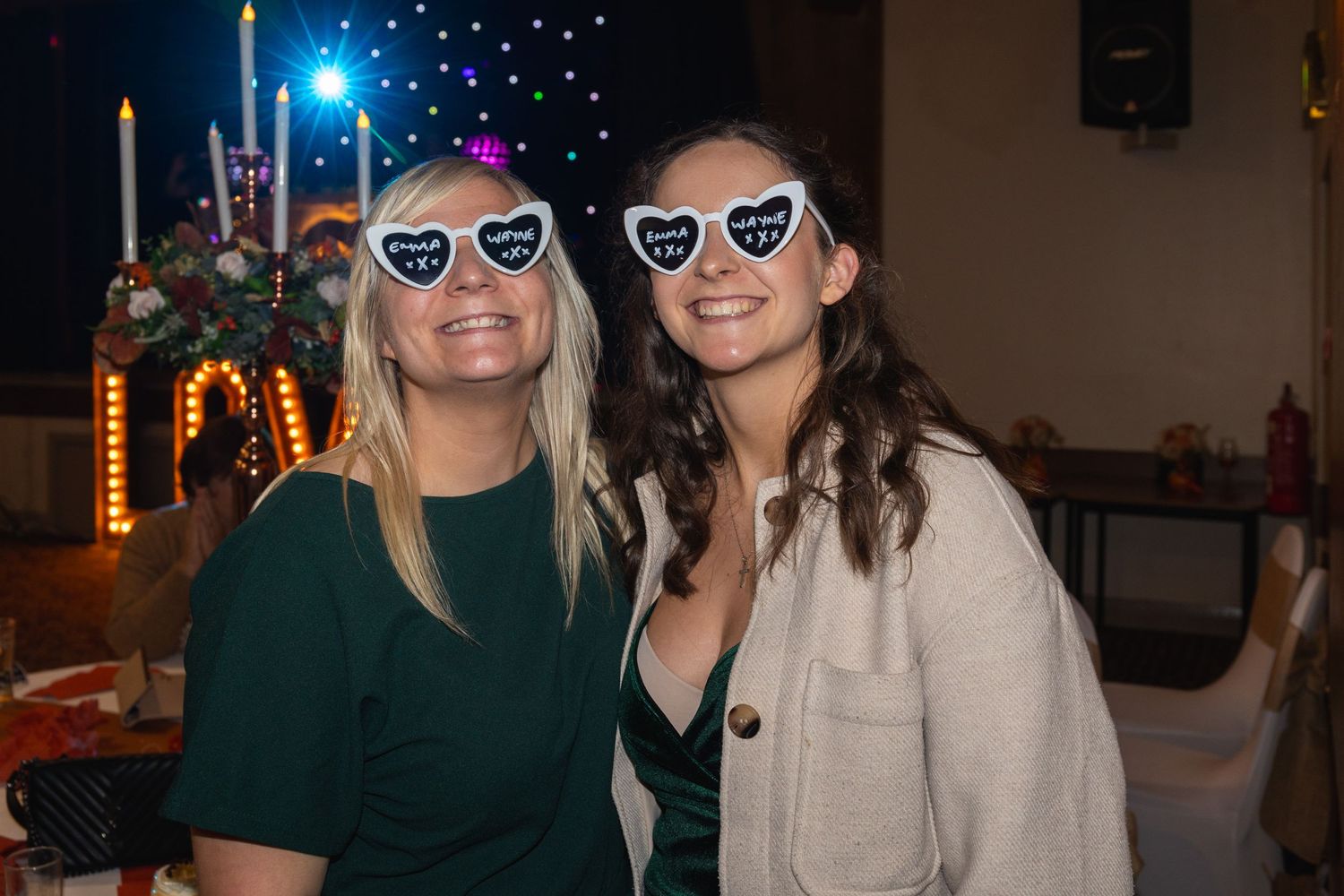 Two people wearing heart-shaped sunglasses pose together at evening event with lit backdrop.