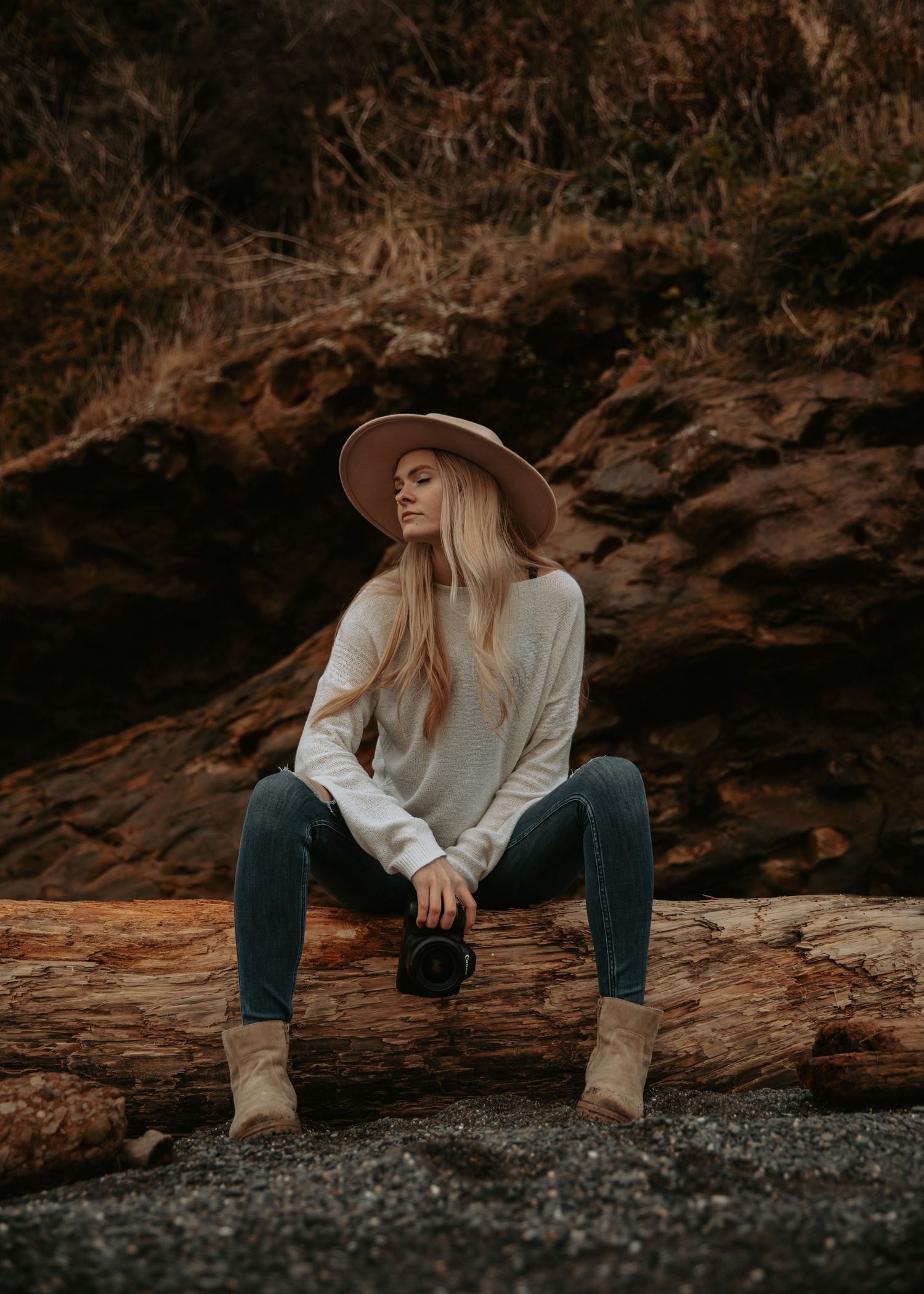 Person in wide brim hat sitting on fallen log against rocky coastal backdrop.