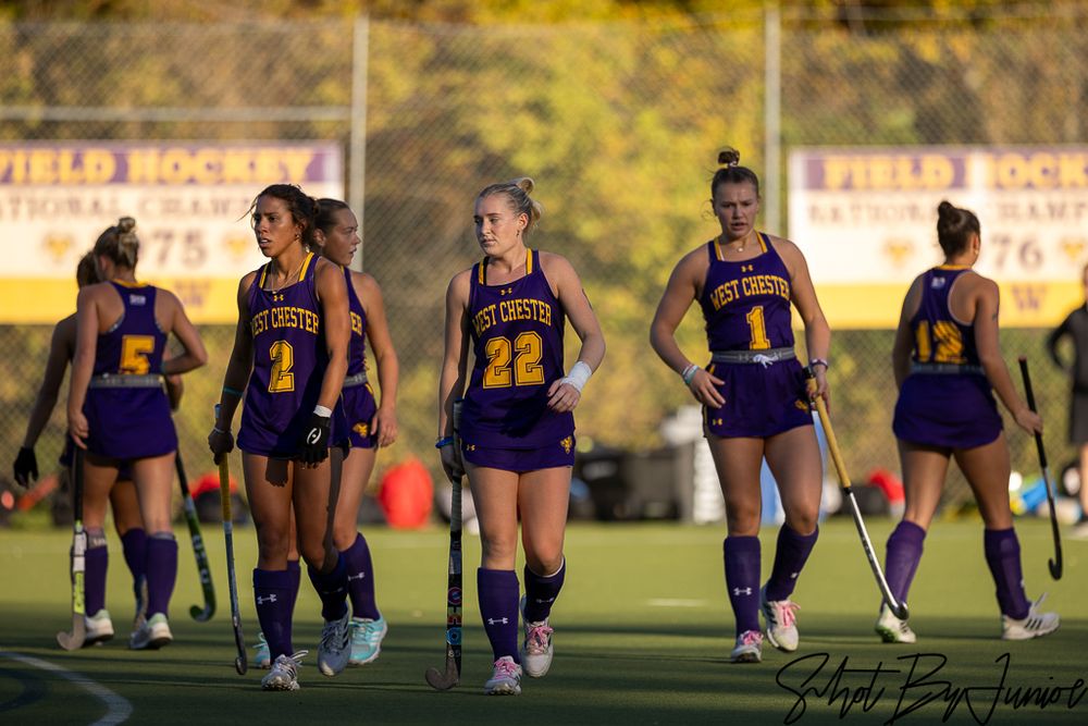 Field hockey team members lined up on the sideline watching their teammates play at dusk.