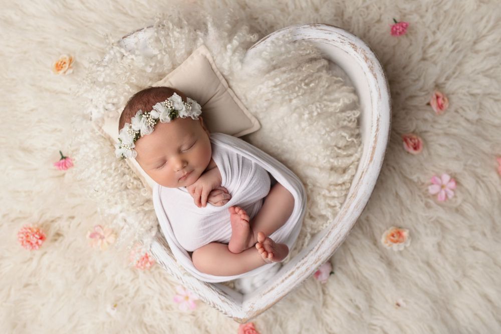 Peaceful newborn in heart-shaped prop with white floral crown and scattered pink petals.
