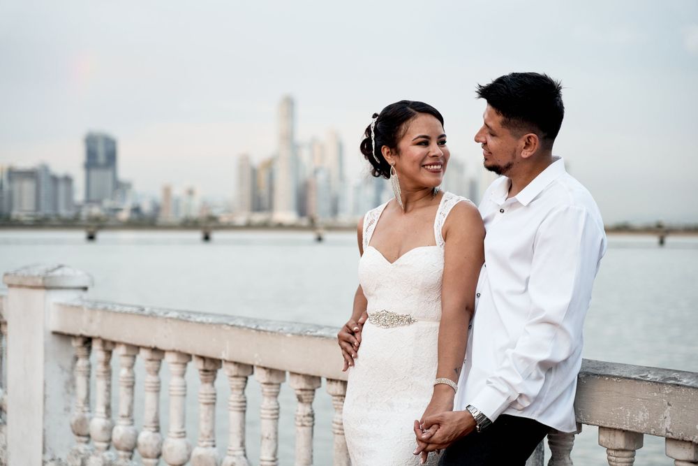 A couple shares an intimate moment along a waterfront balustrade with a city skyline in the background.