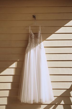 A white lace wedding dress hangs against a tan wall with dramatic window light and shadows.