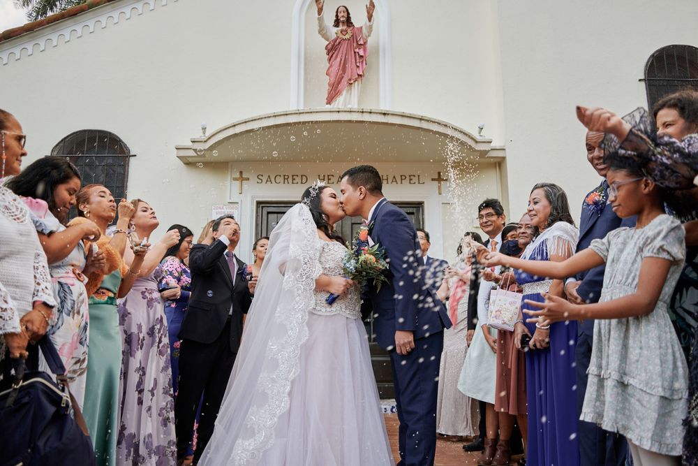 Newlyweds share their first kiss outside a church while guests celebrate with confetti.