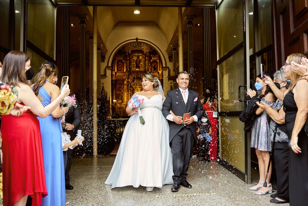 Newlyweds exit a grand church while wedding guests line the sides capturing the moment on their phones.