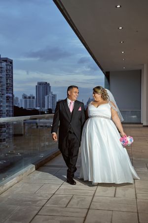 A wedding couple walks together on a modern balcony with city skyline views at dusk.