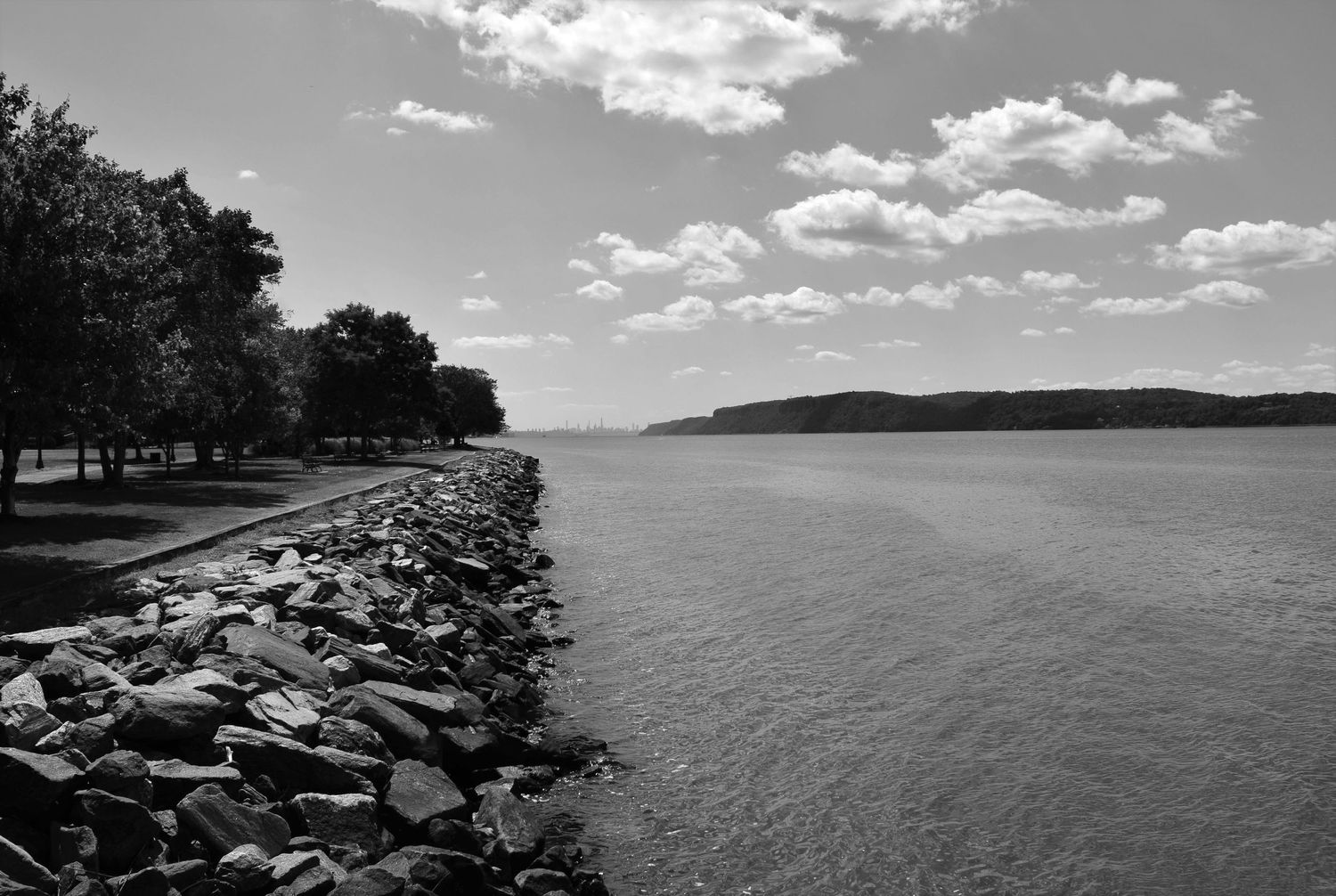 A black and white scenic view of a rocky shoreline along a calm lake with puffy clouds in the sky.