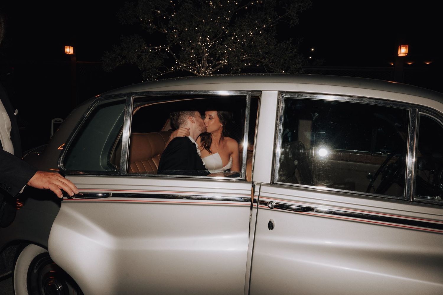 Newlyweds share intimate moment in vintage car during evening departure.