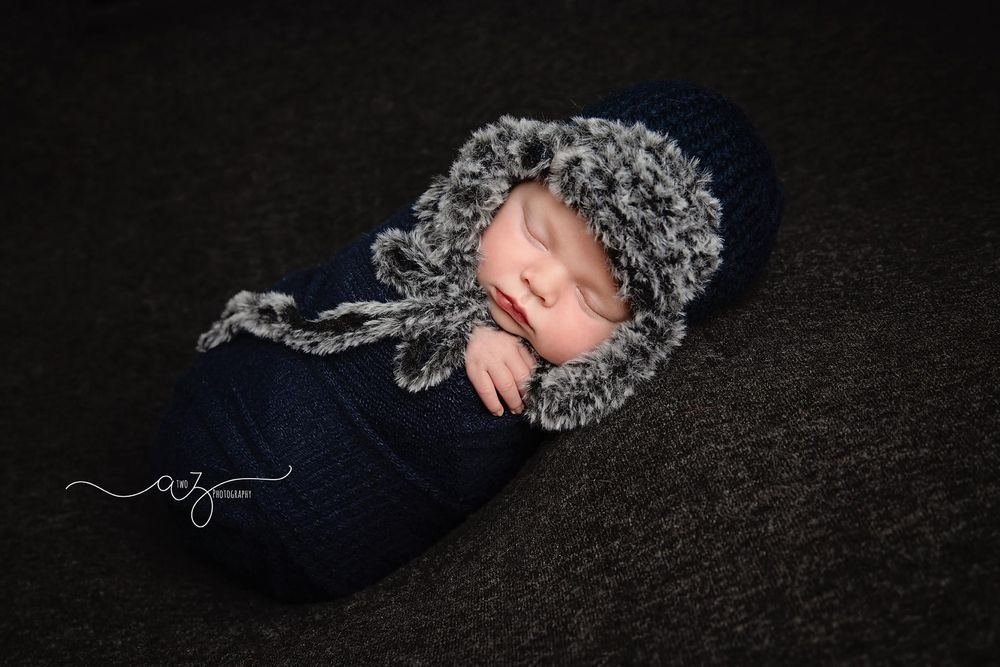 Peaceful sleeping baby in navy outfit and fuzzy winter hat on dark background.