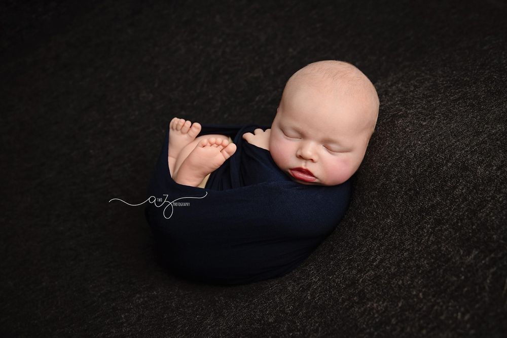 Sleeping newborn swaddled in navy blue wrap against black backdrop.