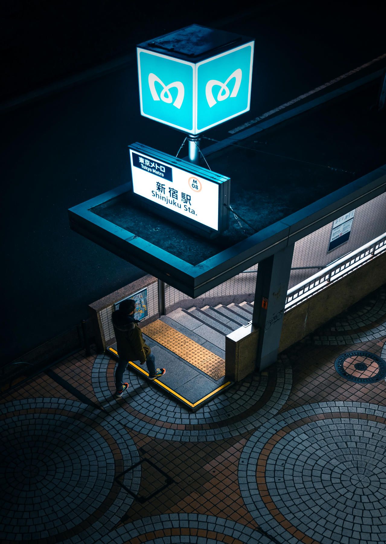 Illuminated blue metro station entrance sign with geometric tiled pavement at night.