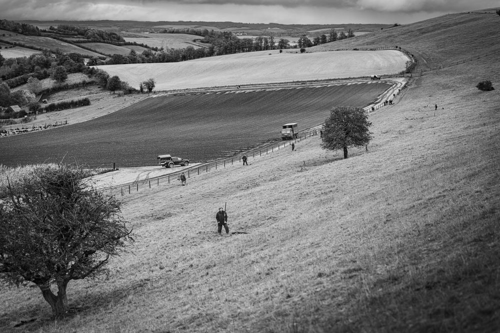 Black and white landscape of rolling hills with scattered trees and hunters in the distance.