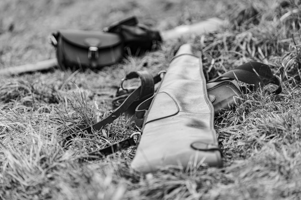 A leather gun case and hunting equipment lying on grass in black and white.