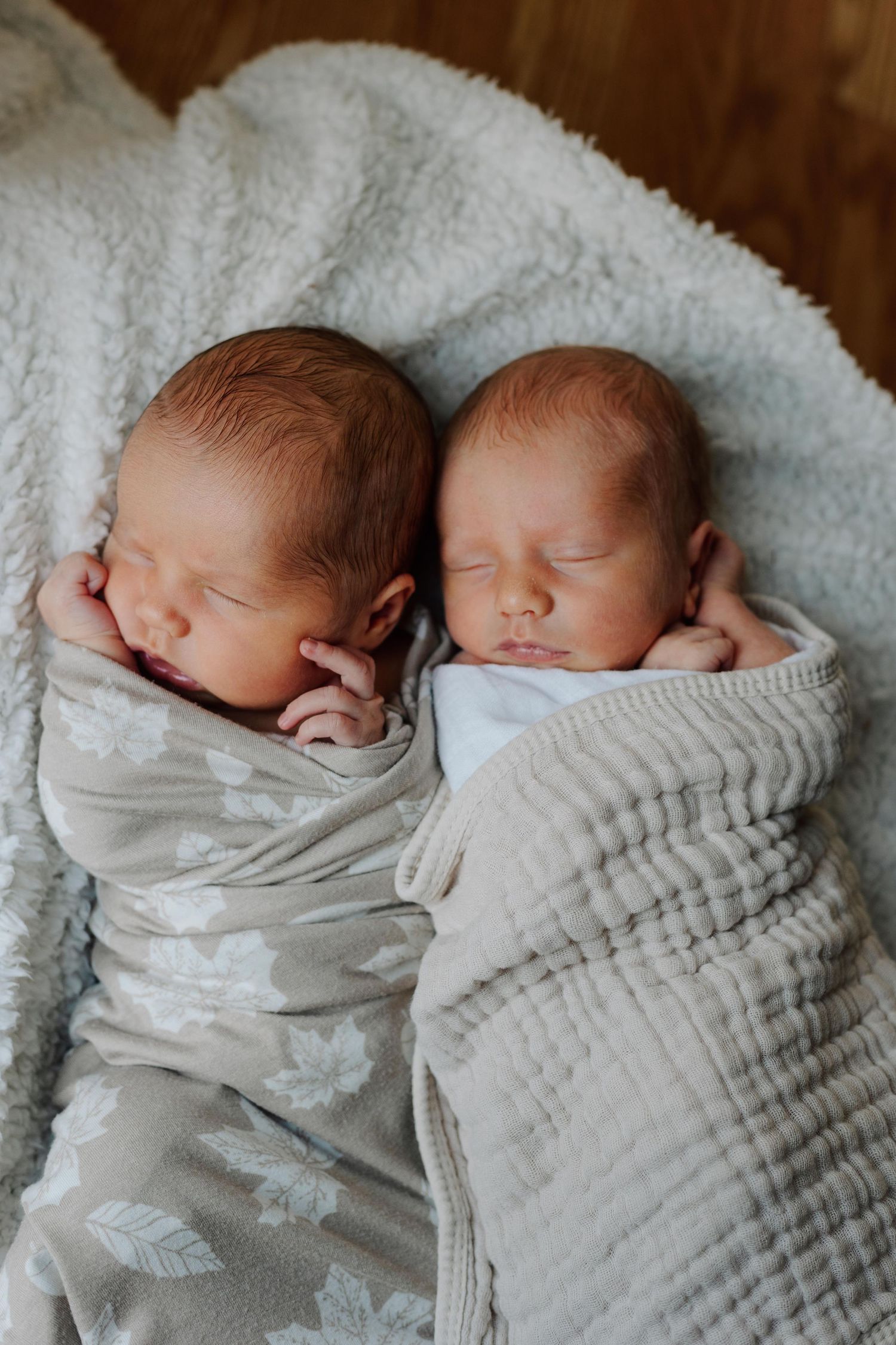 Two sleeping newborn twins swaddled in soft gray blankets on white fluffy fabric.