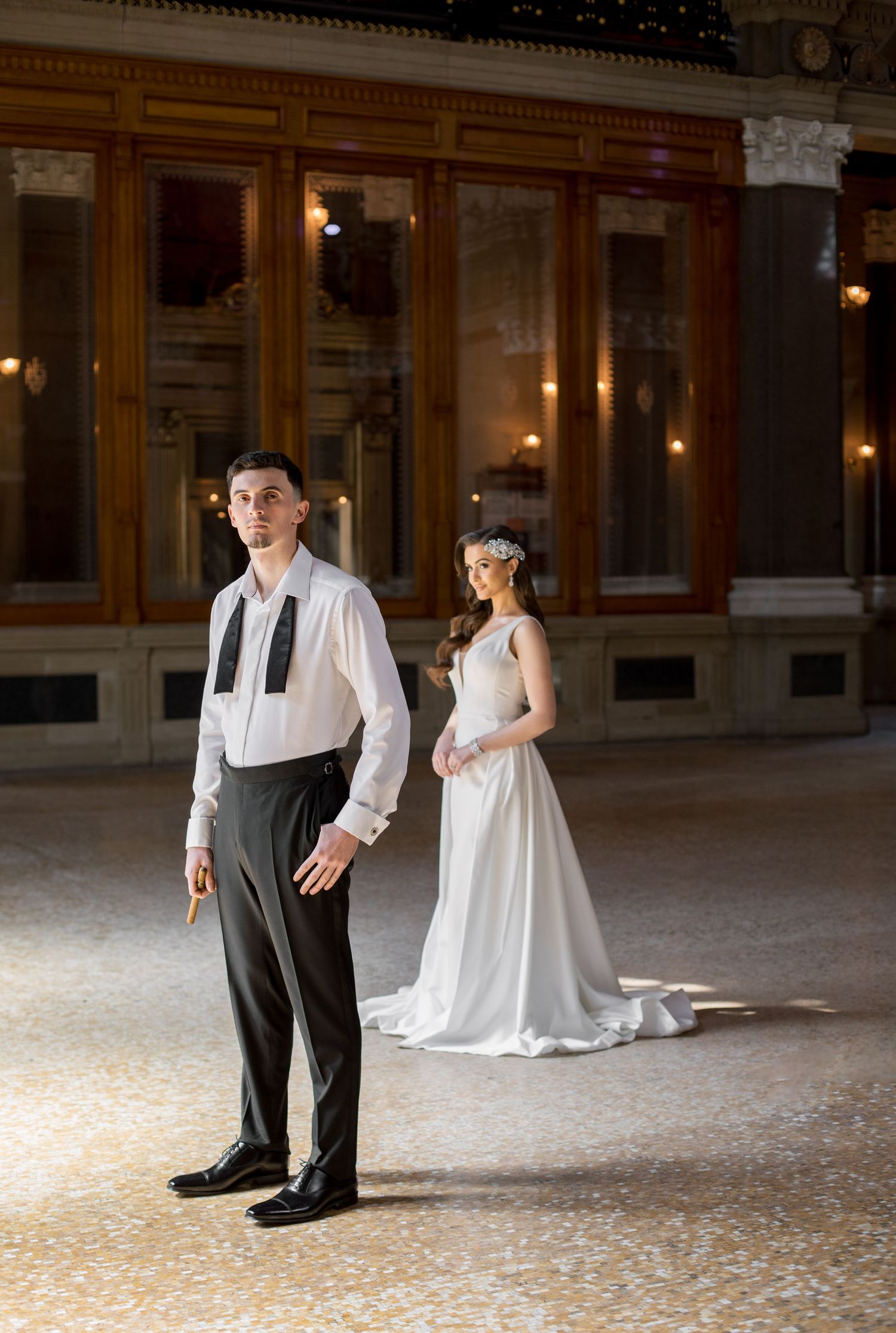 Elegant couple in vintage formal attire standing in a grand historic ballroom.