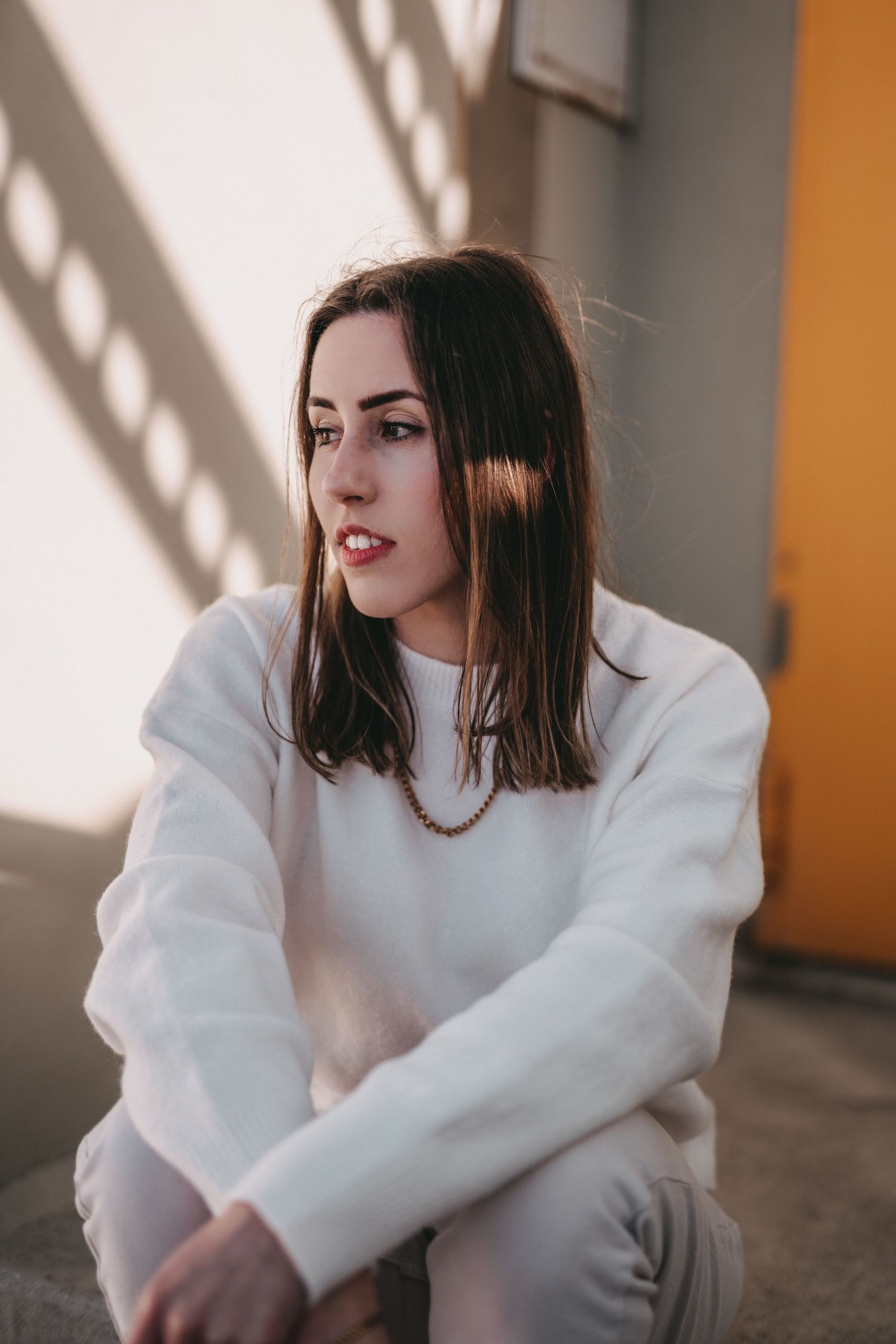 Portrait of someone in a white sweater sitting against a wall with dramatic window light and shadows.
